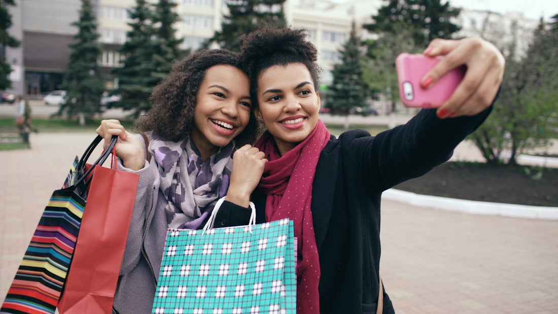 Two smiling women take a selfie with shopping bags.